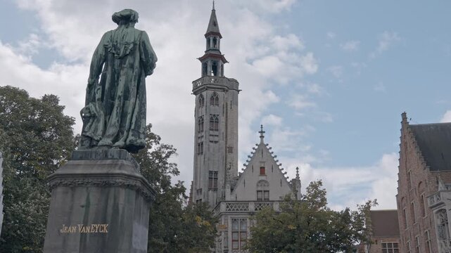 A bronze statue of the Flemish master painter Jan van Eyck stands prominently in Jan van Eyckplein (Jan van Eyck Square) in Bruges, Belgium. In the background, tower of the Poortersloge.