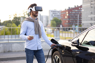 A man wearing a VR headset connects an electric vehicle to a charging station, holding his phone.