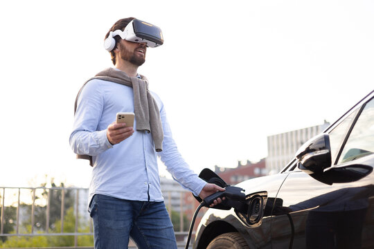 A man wearing a VR headset connects an electric vehicle to a charging station while holding a smartphone. - Powered by Adobe