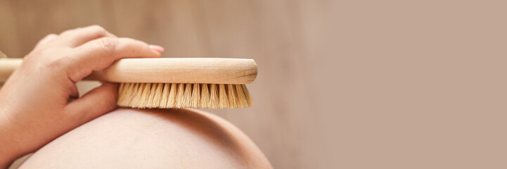 Pregnant female body brushing belly with natural bristle brush for skin care and relaxation.