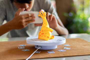 A young person is seen operating a smartphone to capture a series of photographs of a yellow giraffe figurine on a rotating stand for converting the 2D images into a 3D digital model.