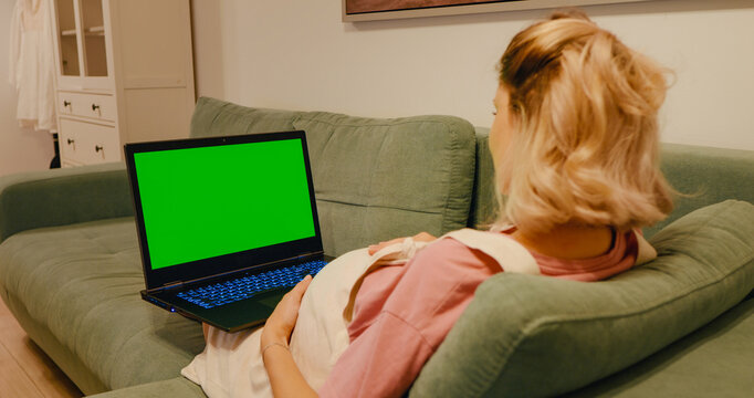 Pregnant looking at green screen copy space laptop while touching her belly. Late pregnancy female having online lessons on the sofa.