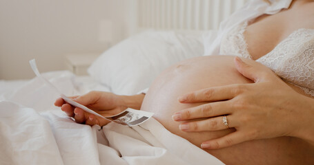 Woman lying in bed admiring ultrasonography picture of her baby and caressing belly. Gravid lady awaits upcoming baby and wants to be good mother.
