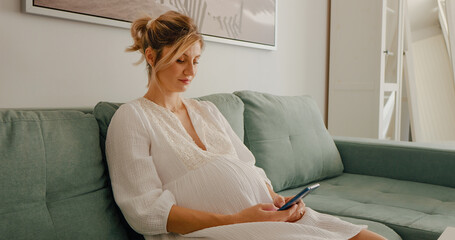 Happy pregnant woman with modern smartphone checking social media on couch at living. Pregnant entertaining herself gaming.