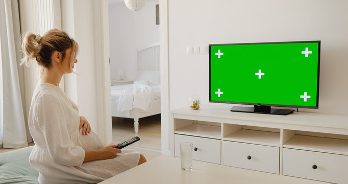 Pregnant woman with remote control watching chroma key tv while sitting on coffee table at home. Side view of woman changing channels.