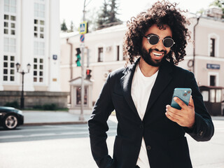 Handsome smiling hipster model.Sexy unshaven Arabian man dressed in summer suit jacket clothes.Fashion male with long curly hairstyle in the street, using smartphone apps, looking at cellphone screen
