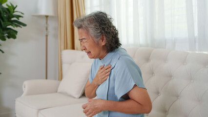 Asian senior woman sitting on sofa touching her chest in pain, feeling discomfort from heart...
