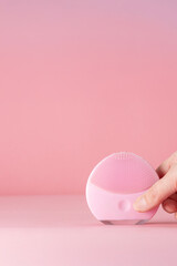 A hand gently holds a pink facial cleansing device on a matching pink background. The minimalist and clean design highlights the product in a sleek, modern, and feminine way.