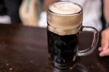 A beautiful, conceptual close-up of a dark beer on the bar counter. The thick head of foam on top suggests the pleasure of drinking. The brewery is in a blurry background, with a hand next to the mug.