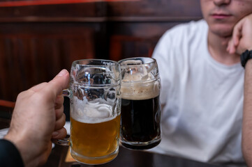 Conceptual shot of friends toasting with a dark and a pale beer at a table. The thick foam on top of the beer hints at the pleasure of drinking. The brewery is in a blurry background.