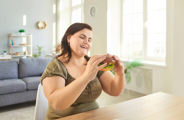 Young fat overweight happy woman sitting at the table in living room at home looking at tasty...