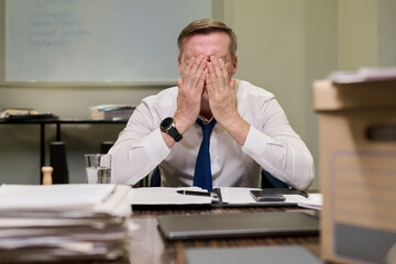 Caucasian middle aged man sitting at desk covering face with hands showing stress or frustration surrounded by paperwork and office supplies in business environment