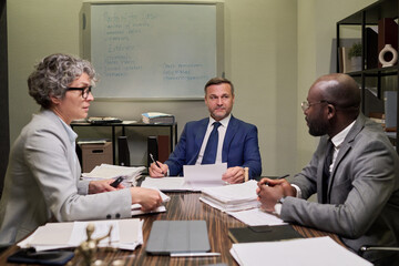 Caucasian middle aged woman, Caucasian middle aged man, Black young man sitting at table discussing documents in office meeting room with paperwork and whiteboard in background