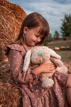 Happy little beautiful child girl hugs and plays with her favorite soft plush toy while sitting on a haystack in a park. Childhood. Autumn background.	