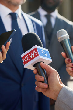 Caucasian middle aged man wearing suit speaking to journalists holding microphones during press conference outdoors, hands of reporters visible in foreground, focus on media interaction