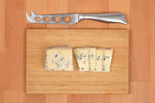 Slices of Gorgonzola blue cheese are placed on a wooden cutting board next to a cheese knife on a wooden surface