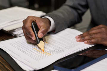 Unrecognizable Black man highlighting text on printed documents with highlighter pen, sitting at desk with smartphone nearby, focusing on reviewing paperwork and marking important information