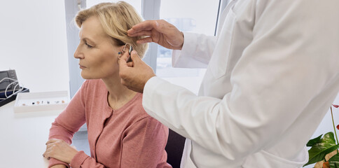 Audiologist putting hearing aid on mature woman's ear at audiology center, close-up, side view. Deafness treatment, hearing solutions