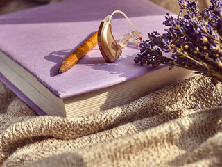 Hearing aid lies next to a pencil on a pastel book, surrounded by lavender and gentle fabric in warm light. Modern technologies for the treatment of hearing