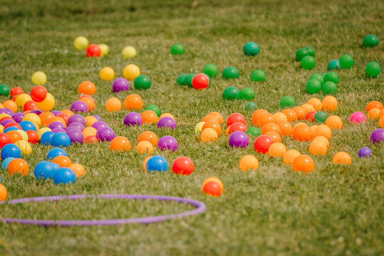 Dozens of colorful plastic balls scattered across a grassy lawn, likely part of an outdoor game or festival activity. Hula hoops visible in the foreground.