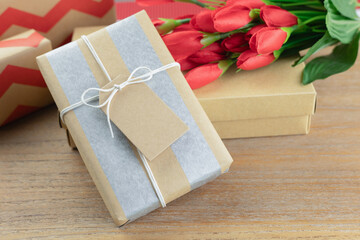 A wrapped gift box with kraft paper and silver stripes, tied with a simple white string and blank tag on a wooden table. and a vibrant bouquet of red tulips, ideal for Valentine's Day, birthdays.