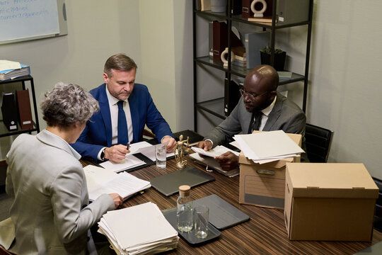 Middle aged Caucasian man and young Black man sitting at table with senior Caucasian woman, reviewing legal documents in office setting with folders and paperwork visible