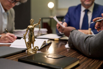 Closeup of Lady Justice statue standing on table with multiethnic group of middle aged men discussing legal documents in office setting, hands holding pens and papers, focus on law theme