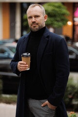Businessman in coat holding paper cup and looking at camera outdoors 