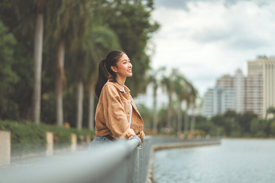 Beautiful Asian woman relaxing by riverside in city park, smiling female enjoying fresh air and peaceful moment, lifestyle and wellness concept with nature, freedom and happiness in urban environment