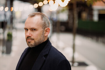 Portrait of elegant man in coat looking at camera on blurred city street