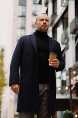 Businessman in coat and turtleneck holding paper cup while walking on street