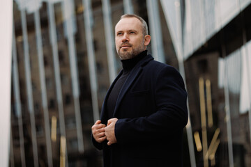 Adult businessman in coat standing near high-rise building on street