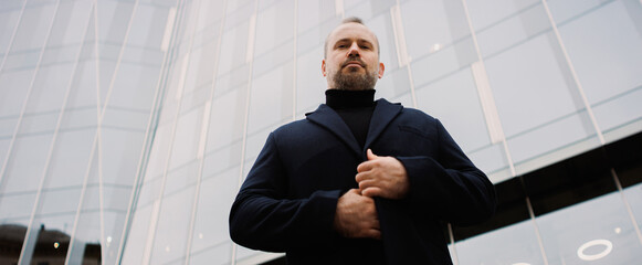 Businessman in coat looking at camera near high-rise building, banner
