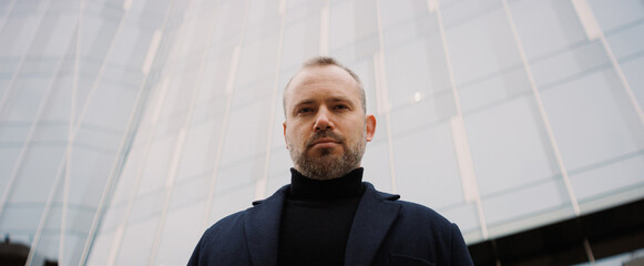 Portrait of businessman in coat looking at camera near high-rise building, banner