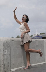 Asian woman poses confidently on a concrete bridge, waving with one hand and smiling in white sunglasses, short skirt, and heels—stylish, playful, and fully engaged with the camera