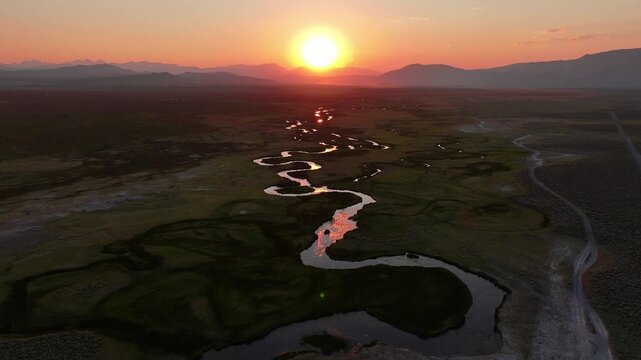 Drone pushes in toward the setting sun over the meandering Owens River near Mammoth Lakes California. Golden hour light glows on S curve channels and open valley with distant Sierra peaks.