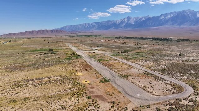 Drone makes a slow parallax orbit around the former Manzanar airfield in Inyo County showing the old runway and taxiway remains set in Owens Valley desert with the Eastern Sierra mountains beyond.