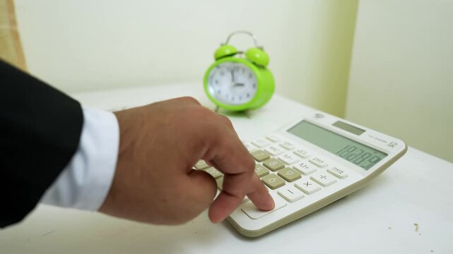 Person in a suit pressing buttons on a calculator with a green alarm clock in the background