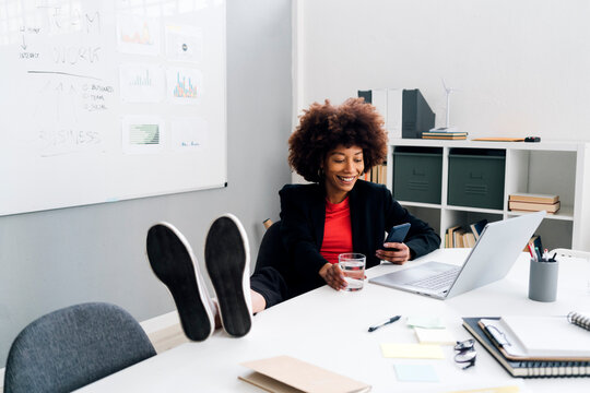 Happy businesswoman sitting with feet up on desk and using mobile phone in office