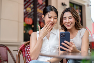 Pretty asian woman showing phone to a friend covering smiling mouth while sits at table outside...