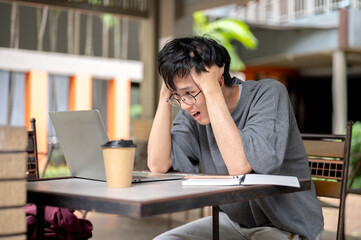 Glasses asian man holding head looking frustrated in front of laptop and book while sitting at table