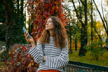 Woman standing in an autumn park, holding a smartphone and looking up, experiencing a thoughtful...