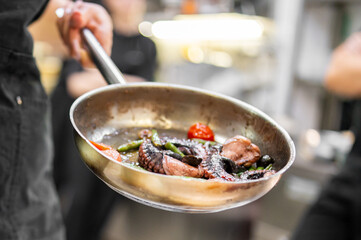 Close-up of gourmet octopus dish with cherry tomatoes, asparagus, and olives in a stainless steel pan, cooked in oil, in a professional kitchen setting.