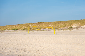 Beach volleyball court. A bright yellow volleyball net stands empty on a sandy beach beneath a clear blue sky. Peaceful coastal dunes stretch in the background.