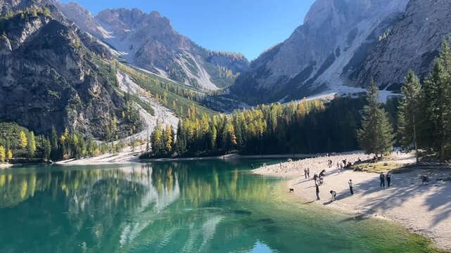 A beautiful panoramic view of Lago di Braies, surrounded by majestic mountains and lush forests, with people enjoying the sandy shores and the clear, reflective waters of the lake.