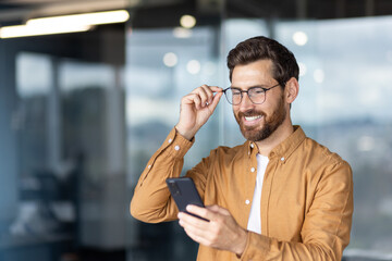 Smiling bearded man adjusting his eyeglasses while holding and interacting with a smartphone,...