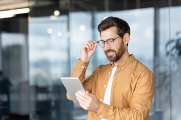 Businessman removes glasses while using a tablet in a bright office, focused on the screen and smiling as he browses data and apps modern workplace technology and innovation