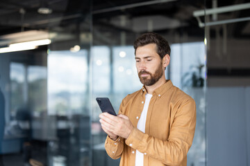 Confident bearded professional standing in a modern office, focused on his smartphone while texting...