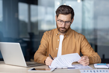 Confident focused businessman working on paperwork while sitting at a desk, reviewing a business...