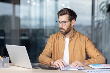 Fototapeta premium Bearded man in glasses focused at a modern office desk, analyzing financial reports and data with a laptop, calculator and paperwork for budgeting and planning